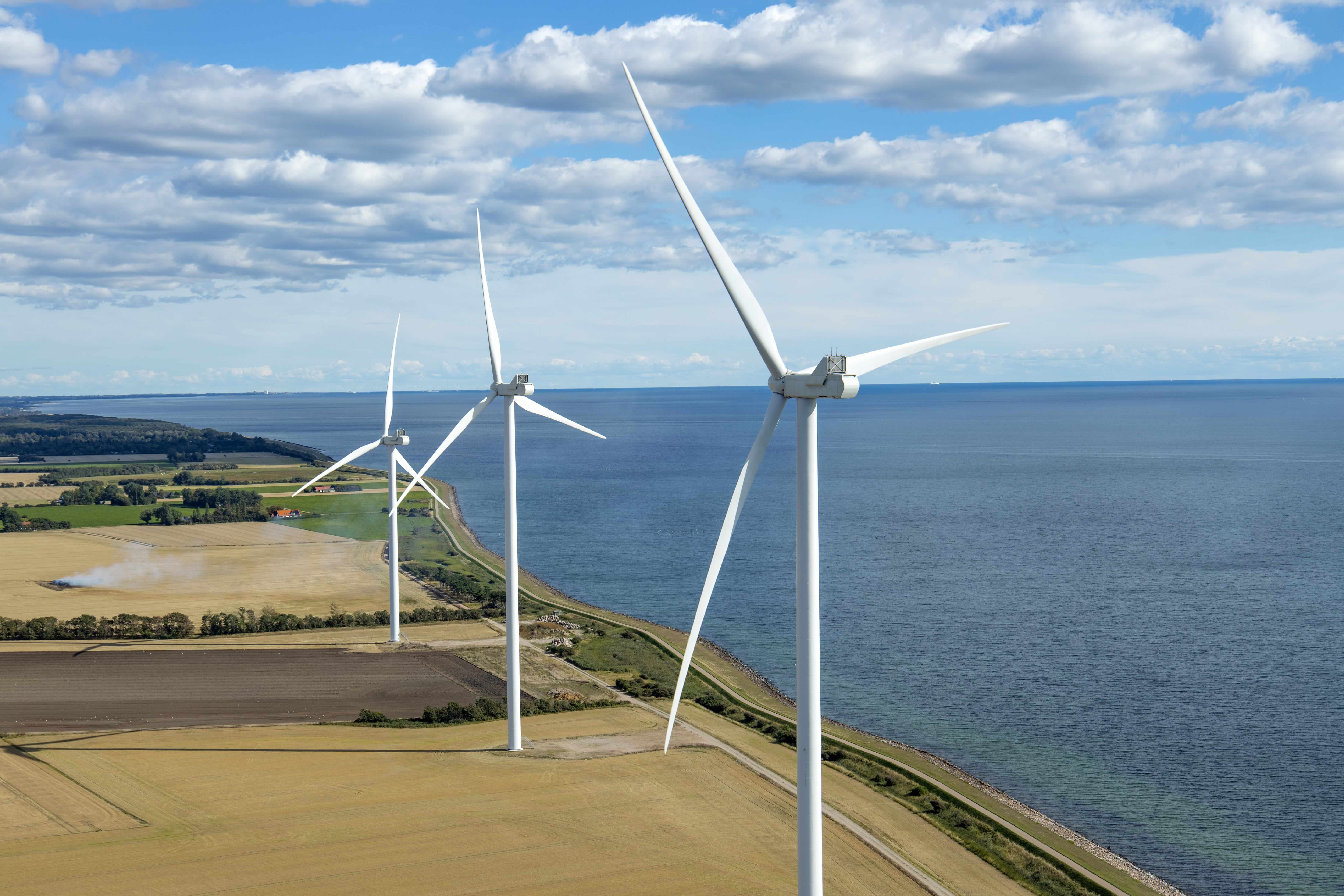 Drei Windräder an einer Küste neben Feldern, im Hintergrund das Meer und ein wolkiger Himmel